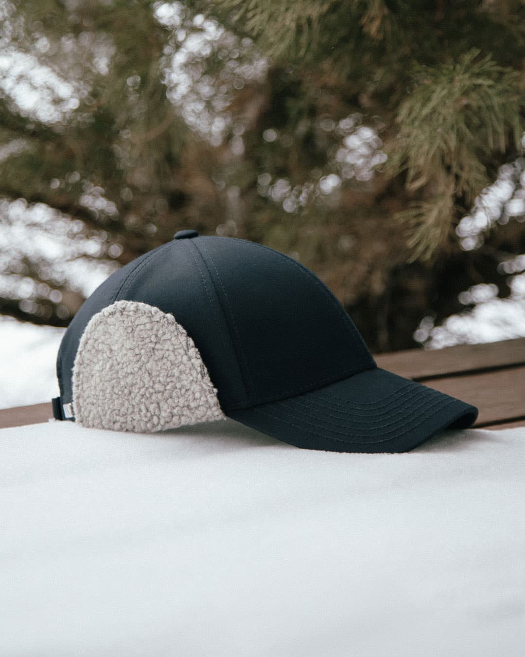 A navy legacy structured cap from Varsity Headwear with teddy ear flaps rests on a snow-covered surface, with blurred trees in the background.