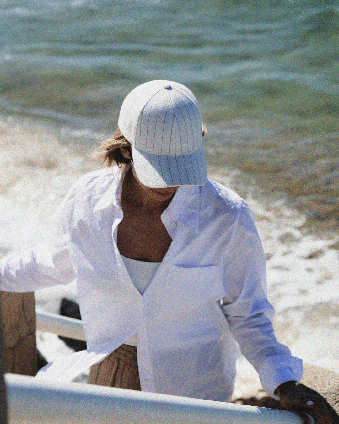 Front view of a limited edition white and beige striped linen cap on a female model at the beach in Cannes