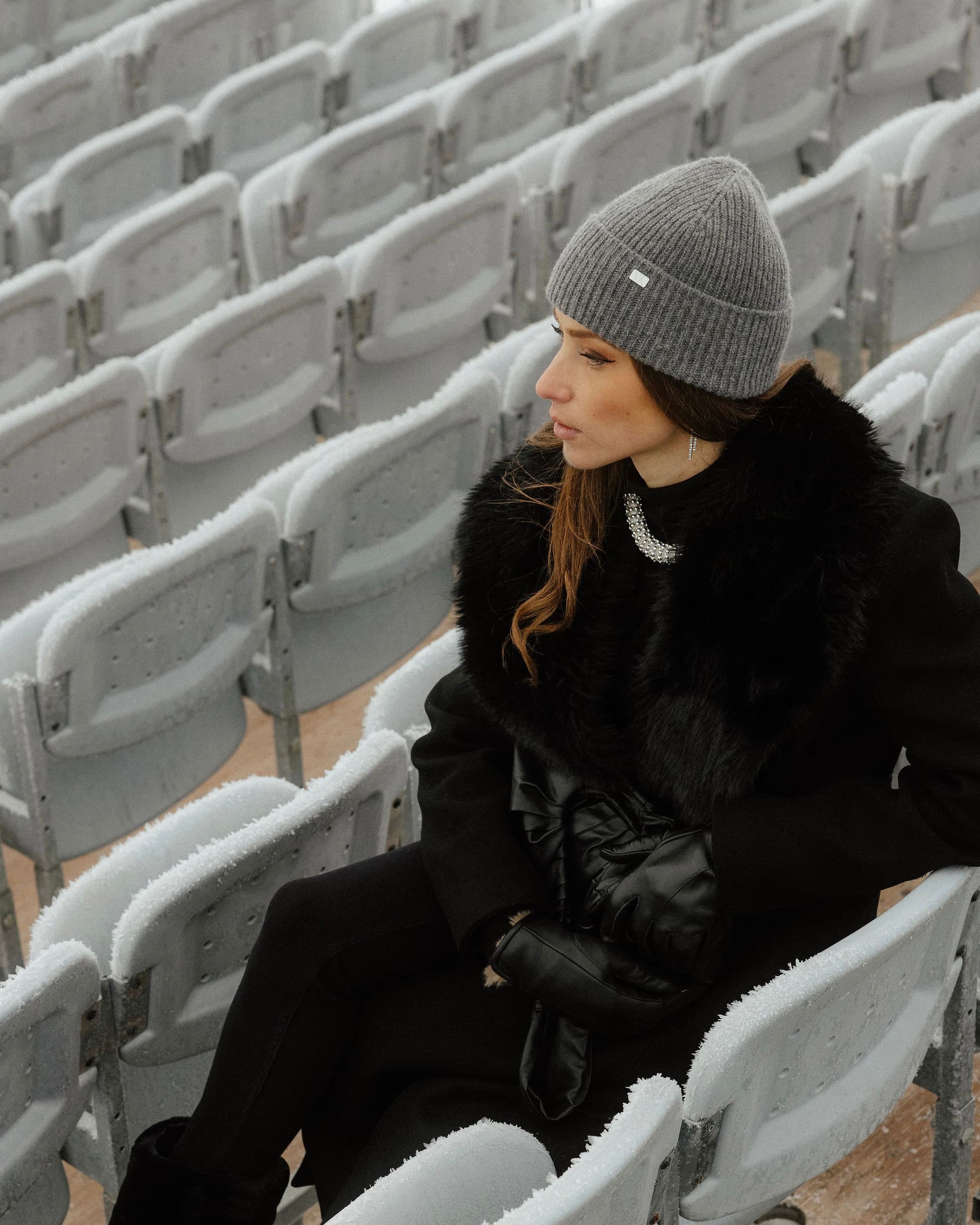 Woman in a black coat and grey beanie from Varsity Headwear sits on frosty stadium seats at The I.C.E in St. Moritz