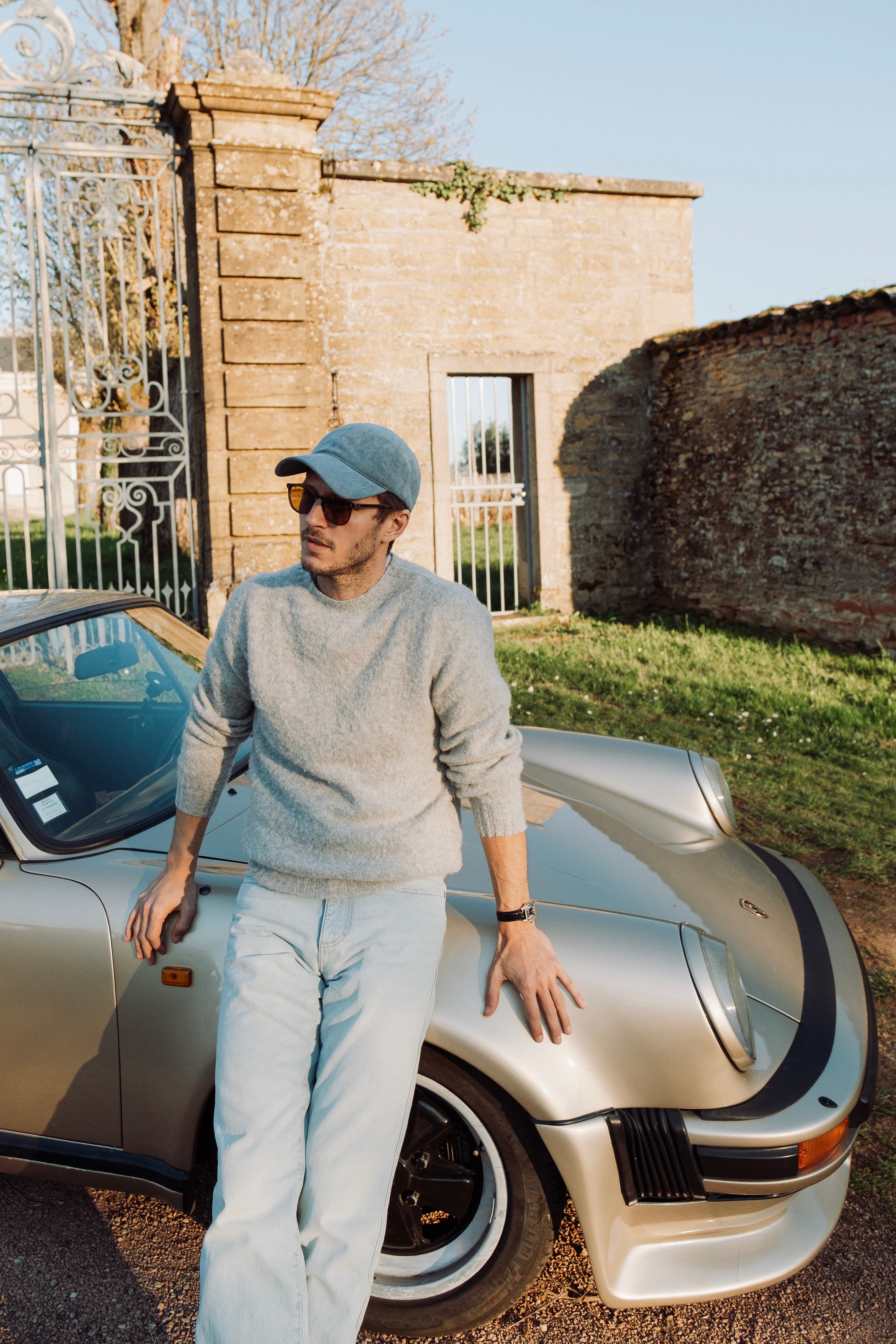 Man in a gray sweater wearing a blue alcantara cap from Varsity Headwear leans against a vintage silver sports car on a sunny day