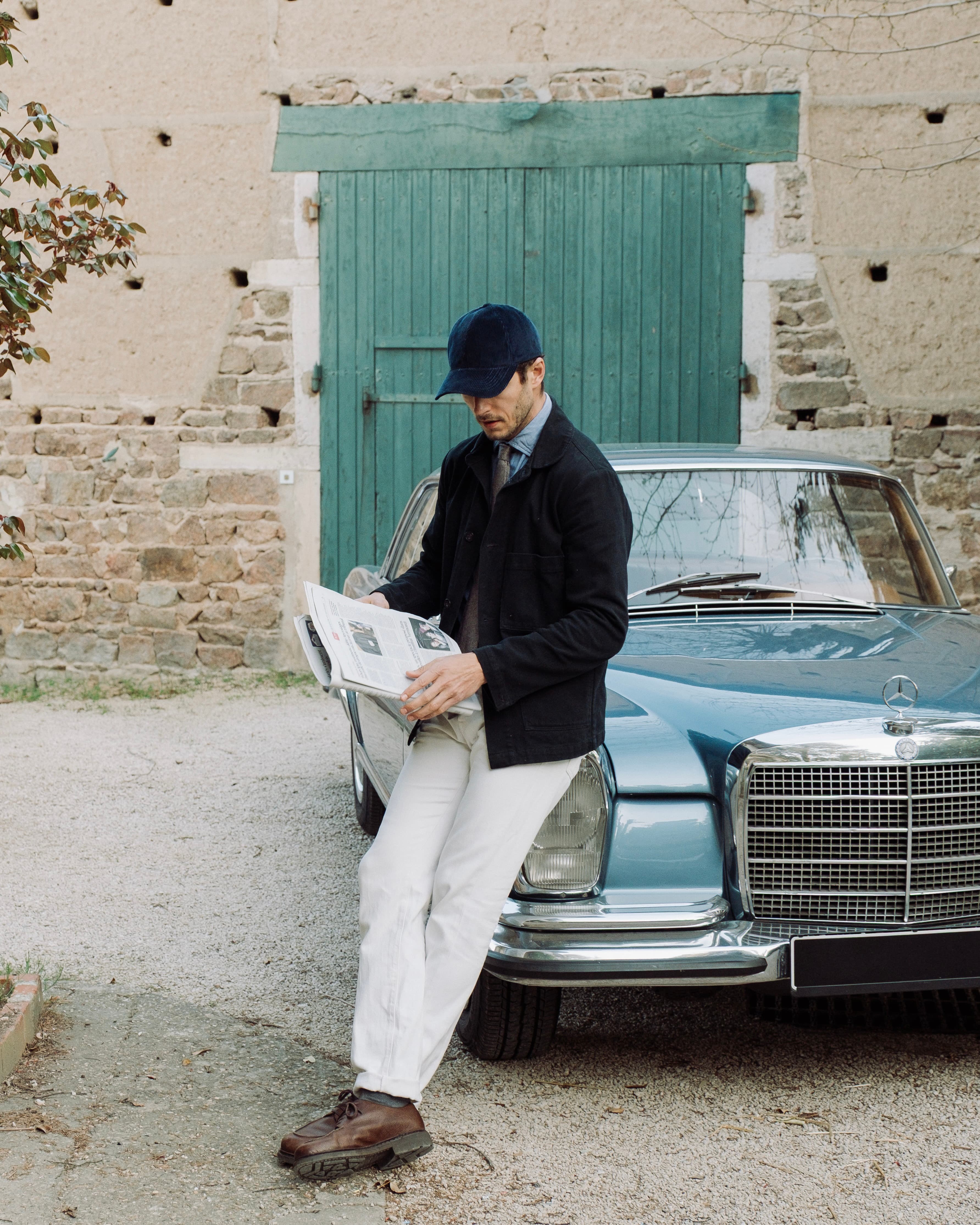 Man in casual attire leans on a vintage blue car, reading a newspaper. Background shows a rustic stone wall with a green wooden door.